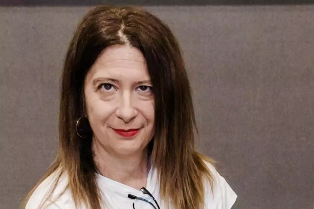 Mujer de cabello castaño lacio y largo, con labios rojos, usando una camiseta blanca y arete de aro, posando frente a un fondo gris. Mujer de cabello castaño lacio y largo, con labios rojos, usando una camiseta blanca y arete de aro, posando frente a un fondo gris.