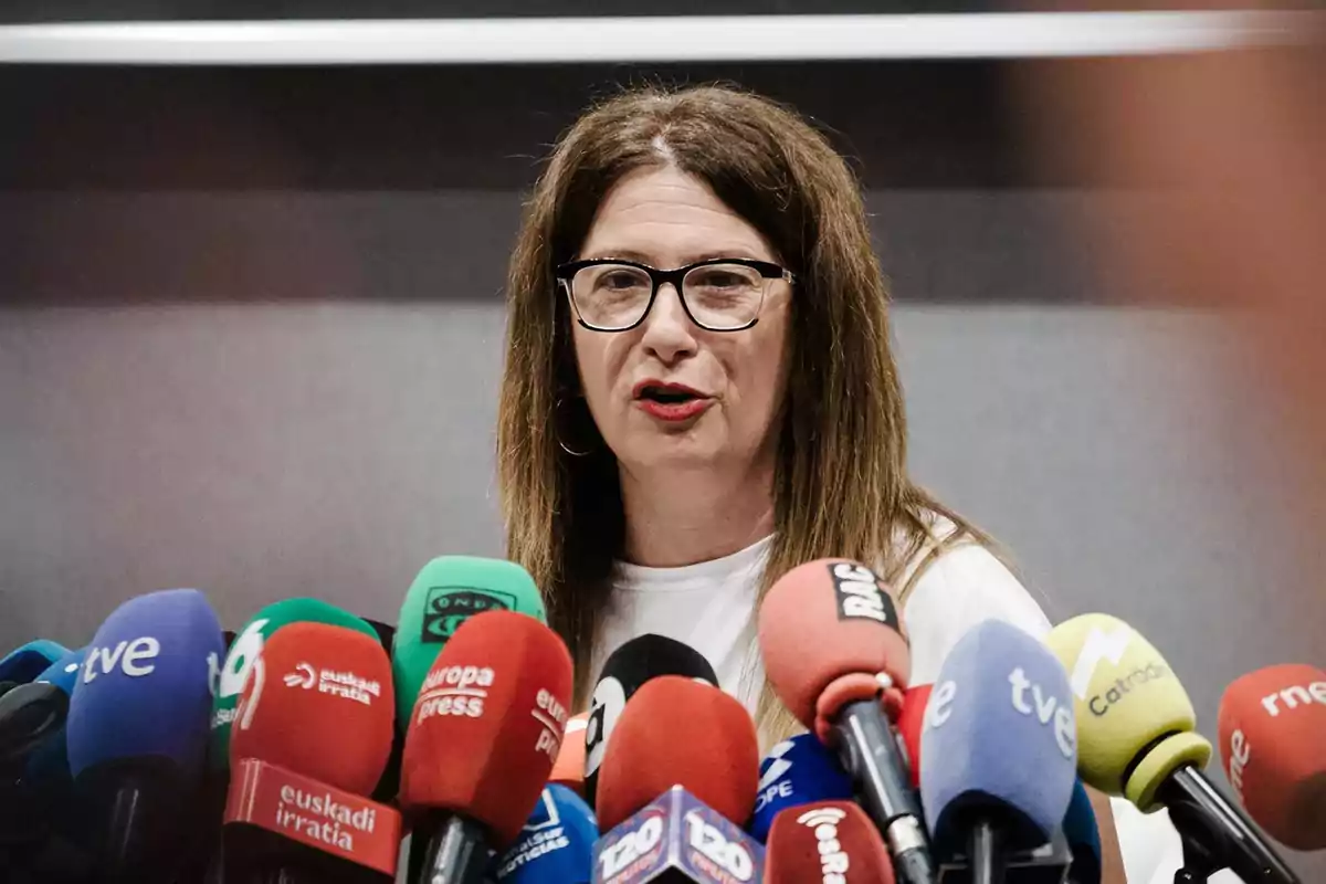 Mujer con gafas hablando frente a numerosos micrófonos de diferentes medios de comunicación