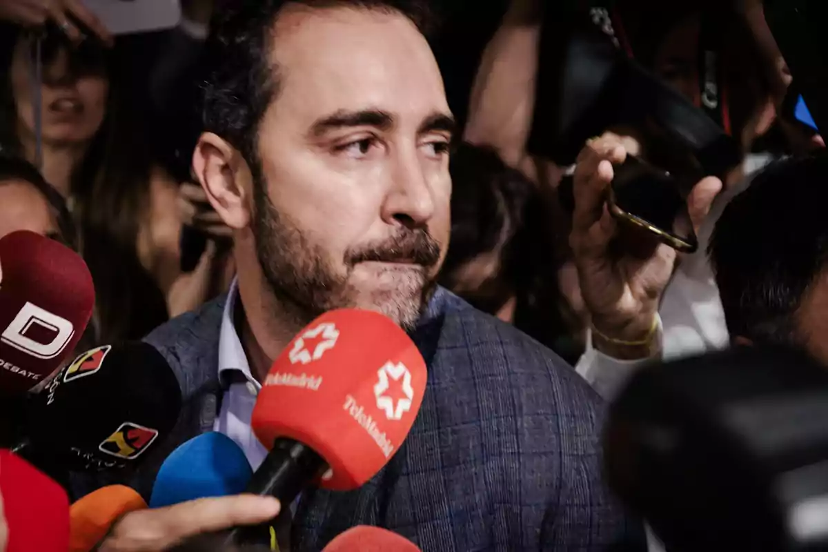 Hombre con barba rodeado de micrófonos y periodistas en una rueda de prensa Hombre con barba rodeado de micrófonos y periodistas en una rueda de prensa
