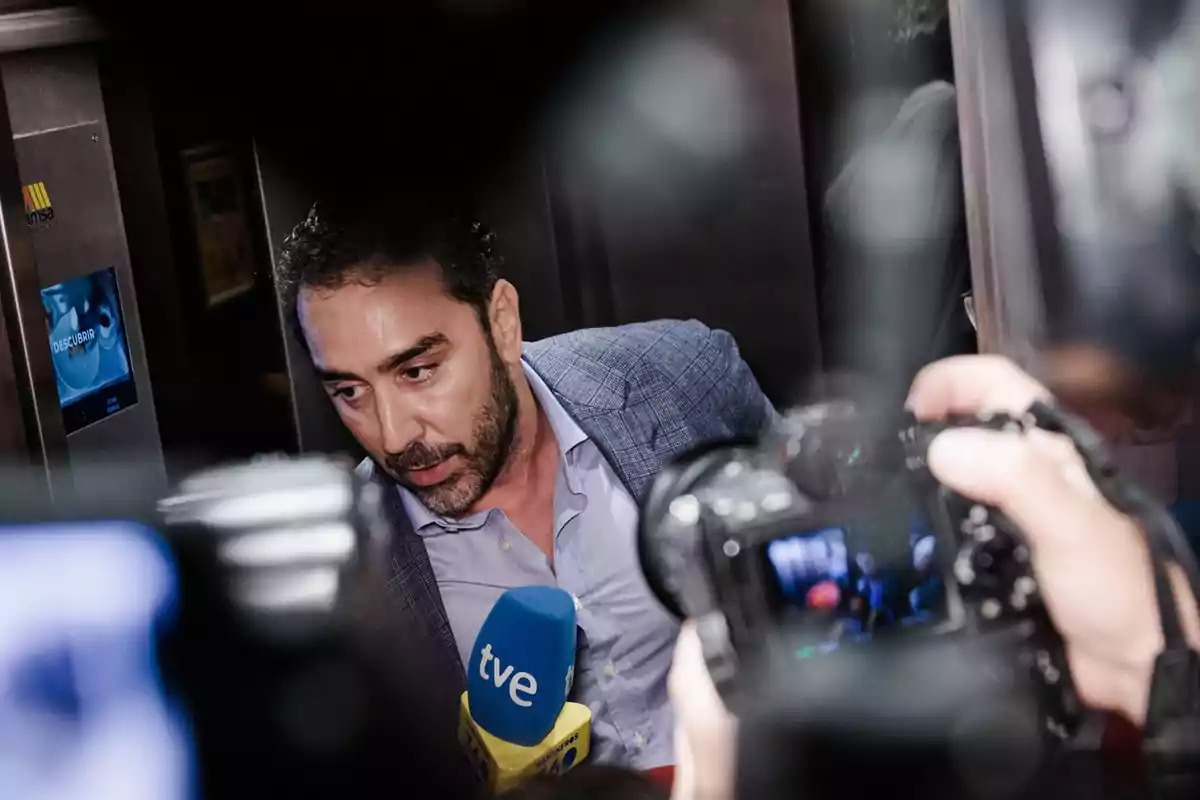 Un hombre con barba y chaqueta azul es entrevistado por varios medios de comunicación frente a un ascensor mientras cámaras y micrófonos lo rodean Un hombre con barba y chaqueta azul es entrevistado por varios medios de comunicación frente a un ascensor mientras cámaras y micrófonos lo rodean