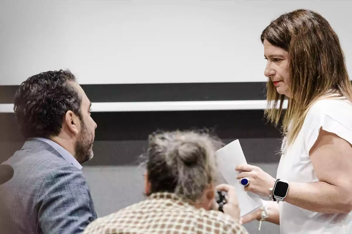 Una mujer de cabello largo sostiene una hoja mientras conversa con un hombre de barba en un entorno de oficina, con otra persona en primer plano tomando una foto.