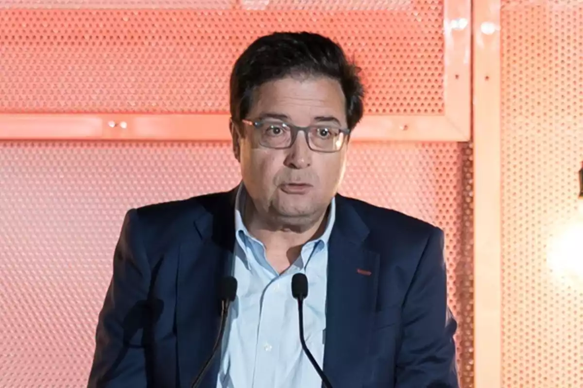 Hombre de mediana edad con gafas y saco azul hablando frente a dos micrófonos con fondo metálico naranja. Hombre de mediana edad con gafas y saco azul hablando frente a dos micrófonos con fondo metálico naranja.