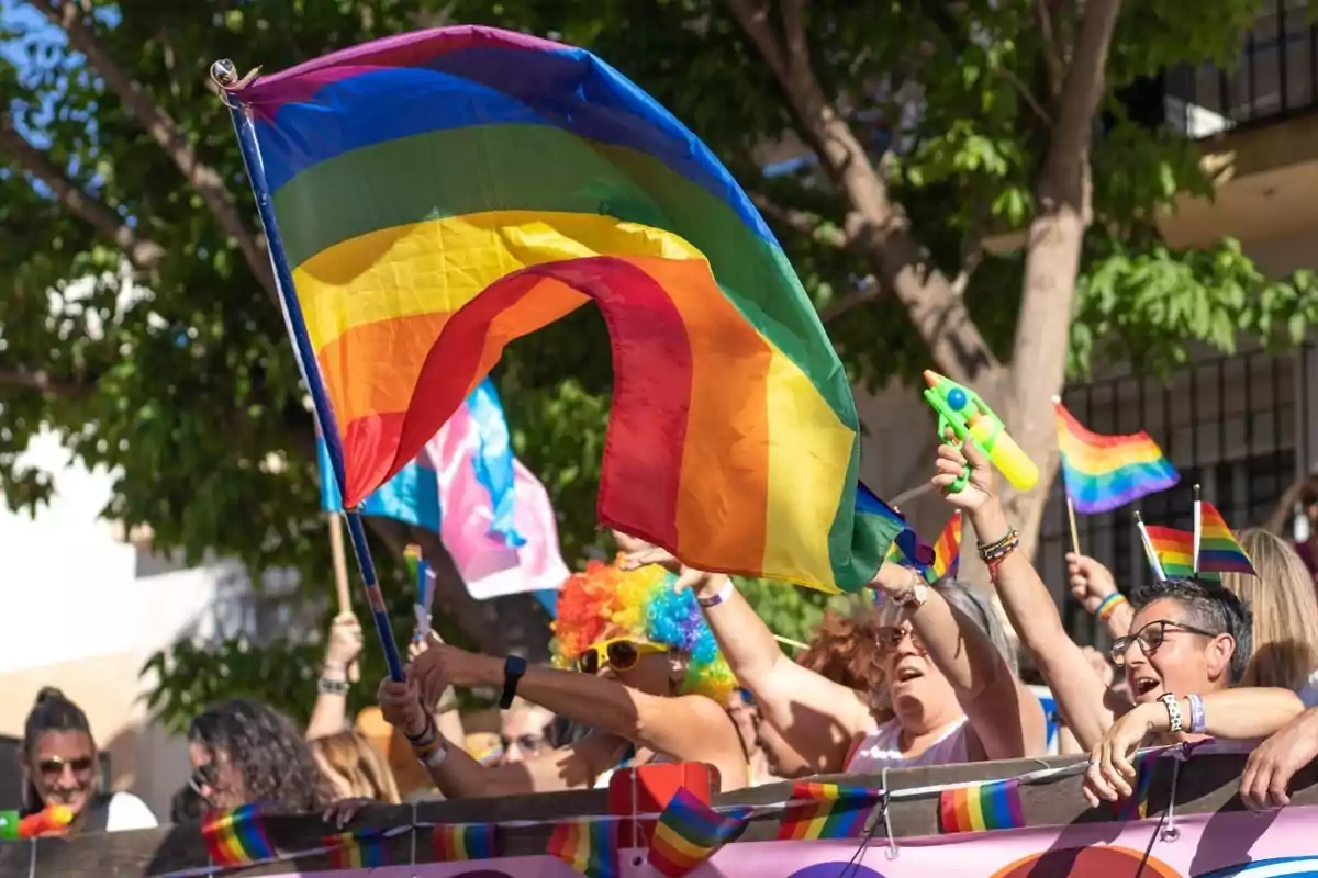Personas celebrando en un desfile del orgullo LGBT con banderas arcoíris y accesorios coloridos bajo árboles en un ambiente festivo Personas celebrando en un desfile del orgullo LGBT con banderas arcoíris y accesorios coloridos bajo árboles en un ambiente festivo