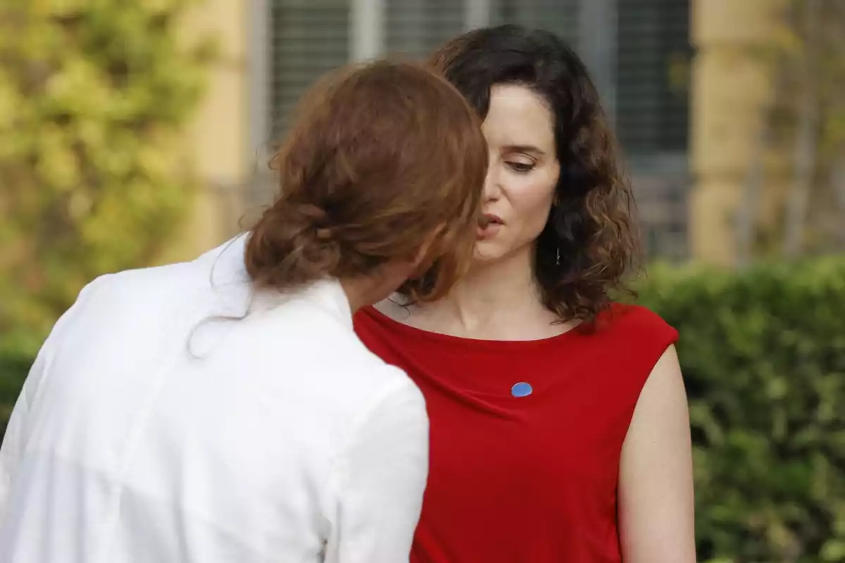 Dos mujeres conversan en un jardín, una de ellas lleva una blusa roja y la otra viste de blanco Dos mujeres conversan en un jardín, una de ellas lleva una blusa roja y la otra viste de blanco