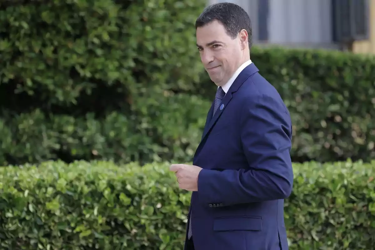 Hombre de cabello corto y oscuro con traje azul caminando al aire libre frente a arbustos verdes Hombre de cabello corto y oscuro con traje azul caminando al aire libre frente a arbustos verdes