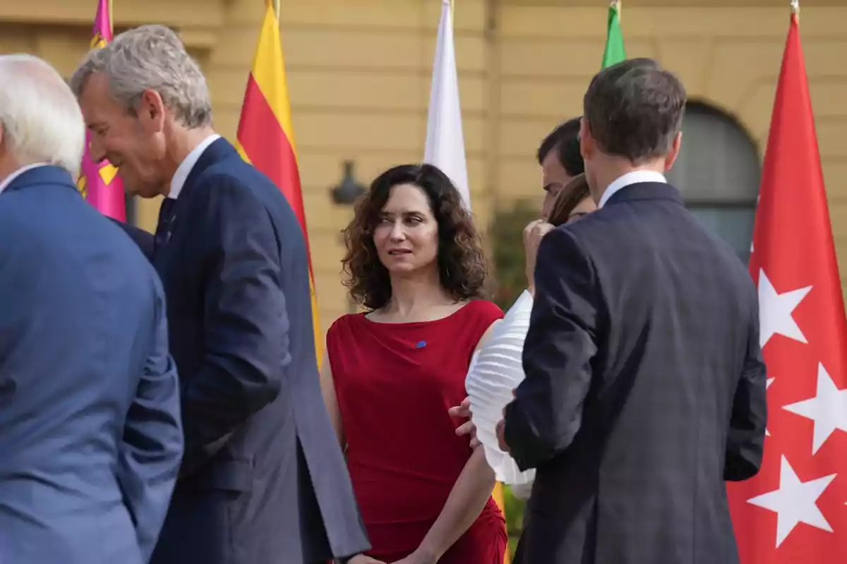 Una mujer con vestido rojo se encuentra entre varias personas vestidas de traje en un evento al aire libre con banderas de fondo Una mujer con vestido rojo se encuentra entre varias personas vestidas de traje en un evento al aire libre con banderas de fondo
