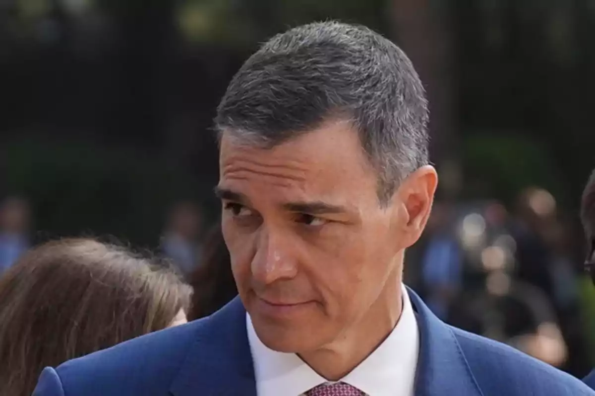 Hombre de cabello corto y canoso con traje azul y corbata rosa mirando hacia un lado en un entorno al aire libre Hombre de cabello corto y canoso con traje azul y corbata rosa mirando hacia un lado en un entorno al aire libre