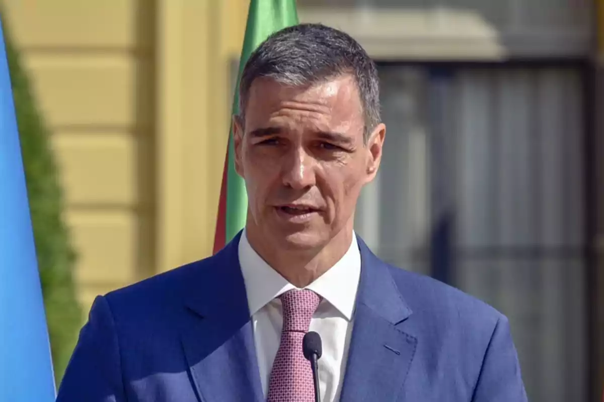 Hombre de cabello corto y canoso con traje azul y corbata rosa hablando frente a un micrófono al aire libre Hombre de cabello corto y canoso con traje azul y corbata rosa hablando frente a un micrófono al aire libre