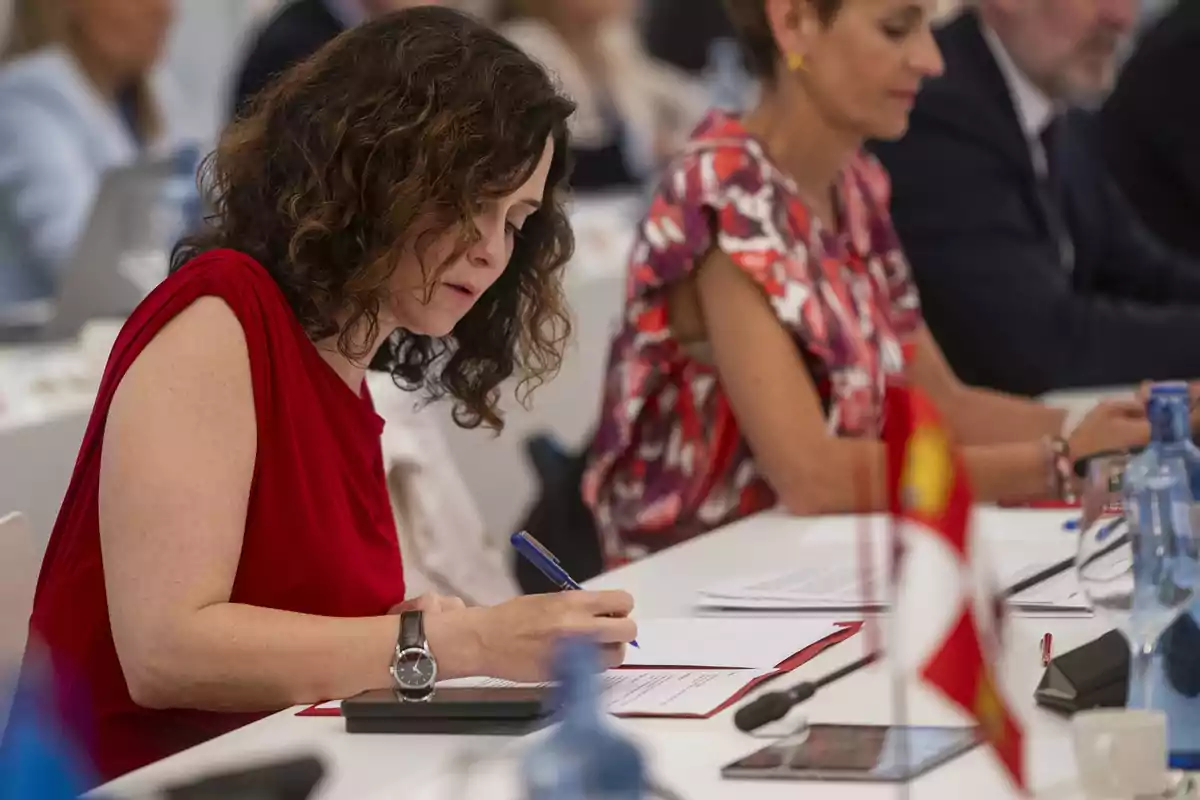 Mujer de cabello rizado y vestido rojo firmando un documento en una mesa de reuniones junto a otras personas Mujer de cabello rizado y vestido rojo firmando un documento en una mesa de reuniones junto a otras personas