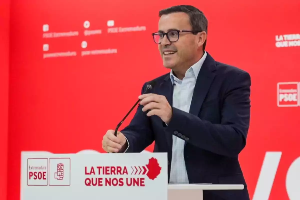 Hombre con gafas y traje oscuro hablando en un atril con fondo rojo y logotipos del PSOE de Extremadura Hombre con gafas y traje oscuro hablando en un atril con fondo rojo y logotipos del PSOE de Extremadura