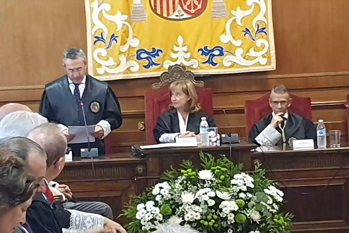 Ceremonia formal en una sala con jueces sentados en una mesa principal y un hombre de pie leyendo un documento, al fondo un tapiz decorativo y en primer plano un arreglo floral blanco y verde. Ceremonia formal en una sala con jueces sentados en una mesa principal y un hombre de pie leyendo un documento, al fondo un tapiz decorativo y en primer plano un arreglo floral blanco y verde.