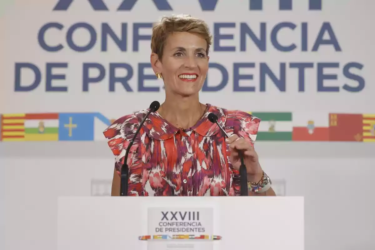 Mujer sonriente con vestido colorido hablando en un podio durante la XXVIII Conferencia de Presidentes con banderas autonómicas de fondo Mujer sonriente con vestido colorido hablando en un podio durante la XXVIII Conferencia de Presidentes con banderas autonómicas de fondo