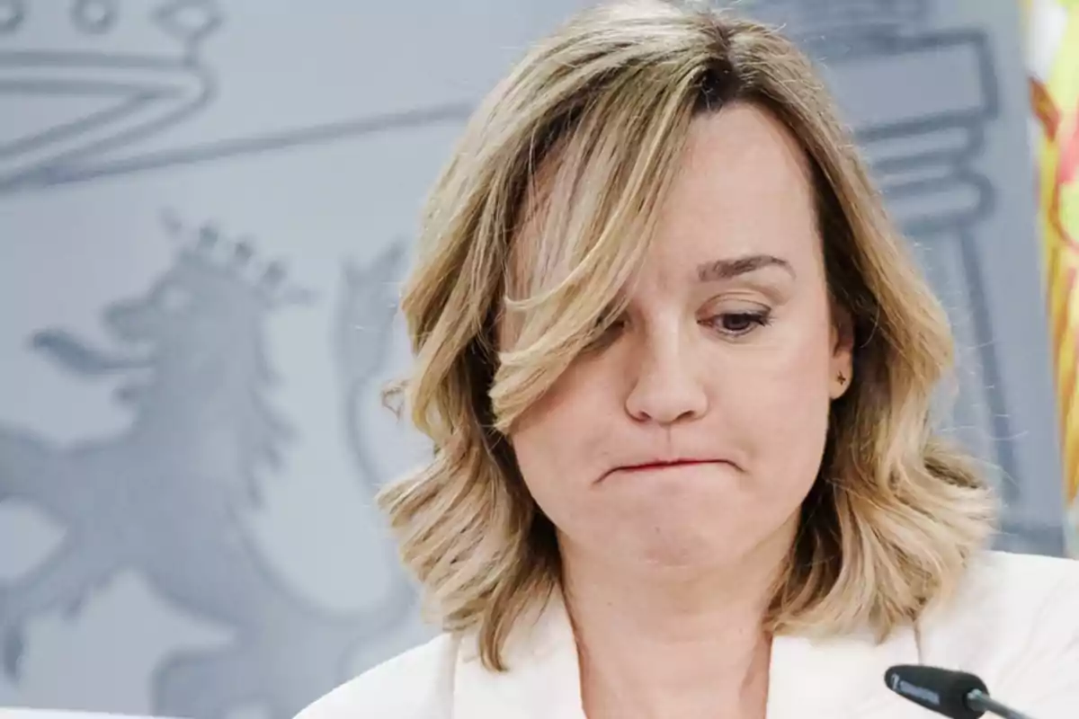 Mujer rubia con expresión seria mirando hacia abajo frente a un fondo institucional Mujer rubia con expresión seria mirando hacia abajo frente a un fondo institucional