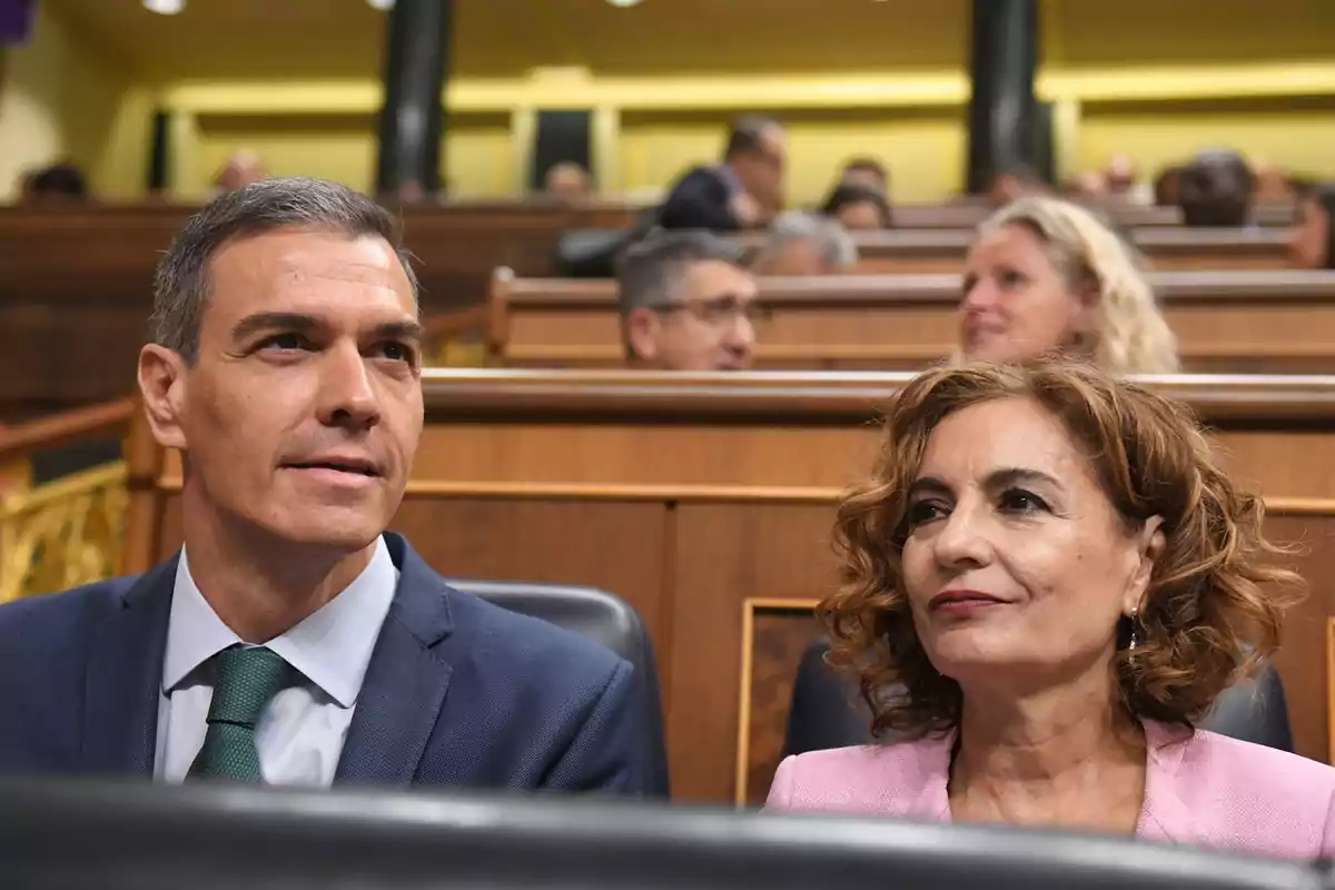 Dos personas sentadas en un recinto parlamentario con otras personas al fondo Dos personas sentadas en un recinto parlamentario con otras personas al fondo