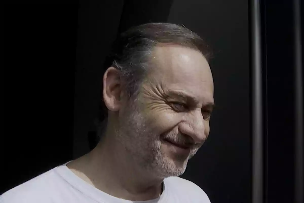 Hombre de mediana edad con barba y cabello corto sonriendo mientras mira hacia abajo en un entorno oscuro Hombre de mediana edad con barba y cabello corto sonriendo mientras mira hacia abajo en un entorno oscuro