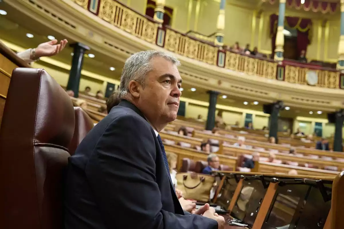 Hombre de cabello canoso sentado en un escaño dentro de un parlamento con otras personas al fondo