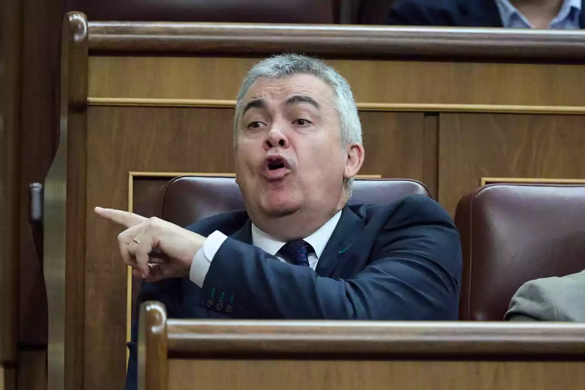 Hombre de cabello canoso con traje oscuro sentado en un escaño señalando con el dedo y hablando en un entorno parlamentario Hombre de cabello canoso con traje oscuro sentado en un escaño señalando con el dedo y hablando en un entorno parlamentario