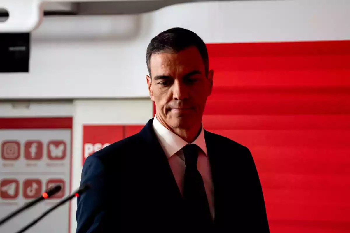 Hombre de traje oscuro y corbata de pie frente a un fondo rojo con logotipos de redes sociales al fondo Hombre de traje oscuro y corbata de pie frente a un fondo rojo con logotipos de redes sociales al fondo