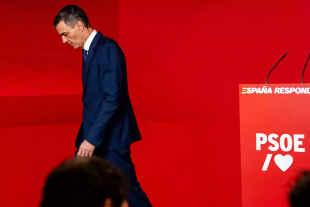 Hombre con traje azul caminando cabizbajo junto a un atril rojo con el logo de PSOE y un fondo rojo Hombre con traje azul caminando cabizbajo junto a un atril rojo con el logo de PSOE y un fondo rojo