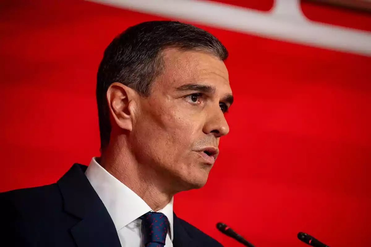 Hombre de cabello corto y canoso hablando frente a micrófonos con fondo rojo