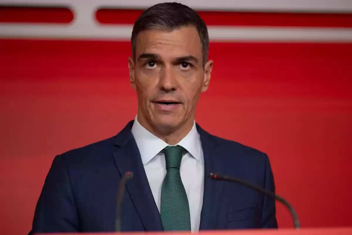 Hombre de cabello corto y canoso con traje azul y corbata verde hablando frente a micrófonos sobre un fondo rojo Hombre de cabello corto y canoso con traje azul y corbata verde hablando frente a micrófonos sobre un fondo rojo