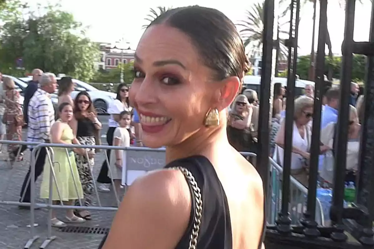 Eva González sonriente con vestido elegante y pendientes dorados en un evento al aire libre con varias personas al fondo Eva González sonriente con vestido elegante y pendientes dorados en un evento al aire libre con varias personas al fondo