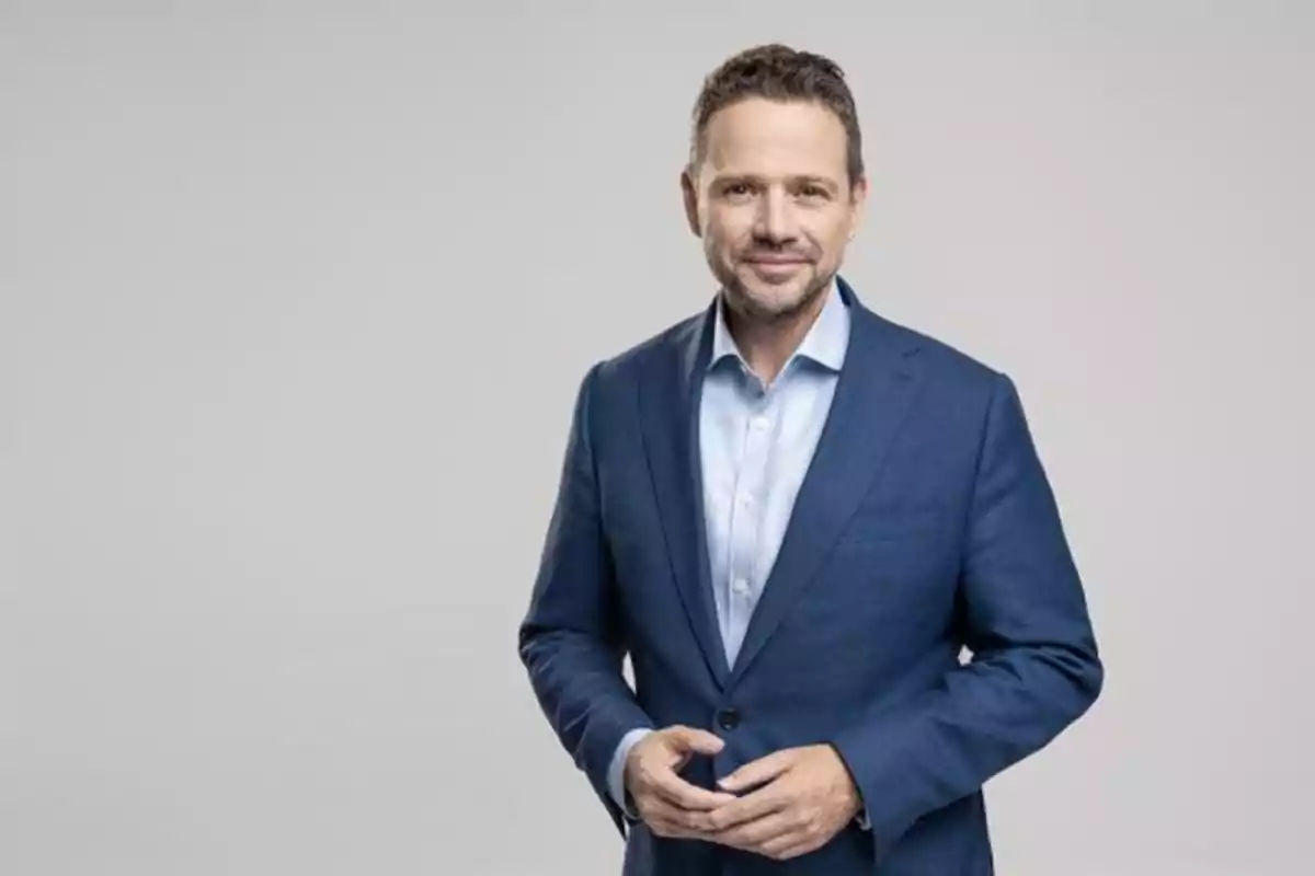 Hombre de mediana edad con barba corta y cabello oscuro viste un traje azul y camisa clara posando frente a un fondo gris claro Hombre de mediana edad con barba corta y cabello oscuro viste un traje azul y camisa clara posando frente a un fondo gris claro