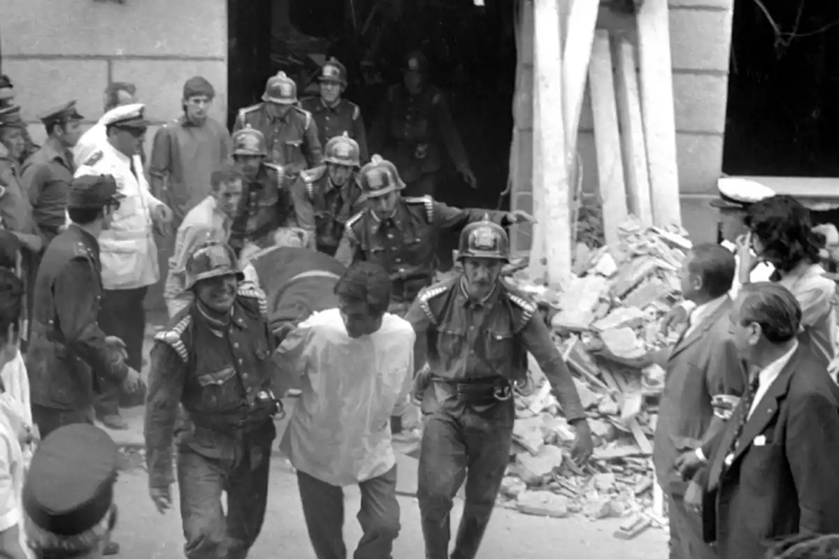 Un grupo de bomberos y personas en uniforme ayudan a evacuar a un hombre de un edificio dañado con escombros alrededor.