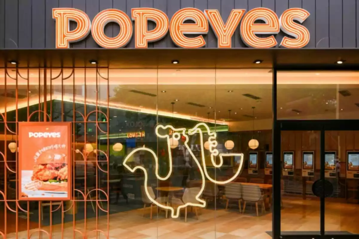 Fachada de un restaurante Popeyes con un letrero de neón en forma de gallo y un cartel de comida en la entrada. Fachada de un restaurante Popeyes con un letrero de neón en forma de gallo y un cartel de comida en la entrada.