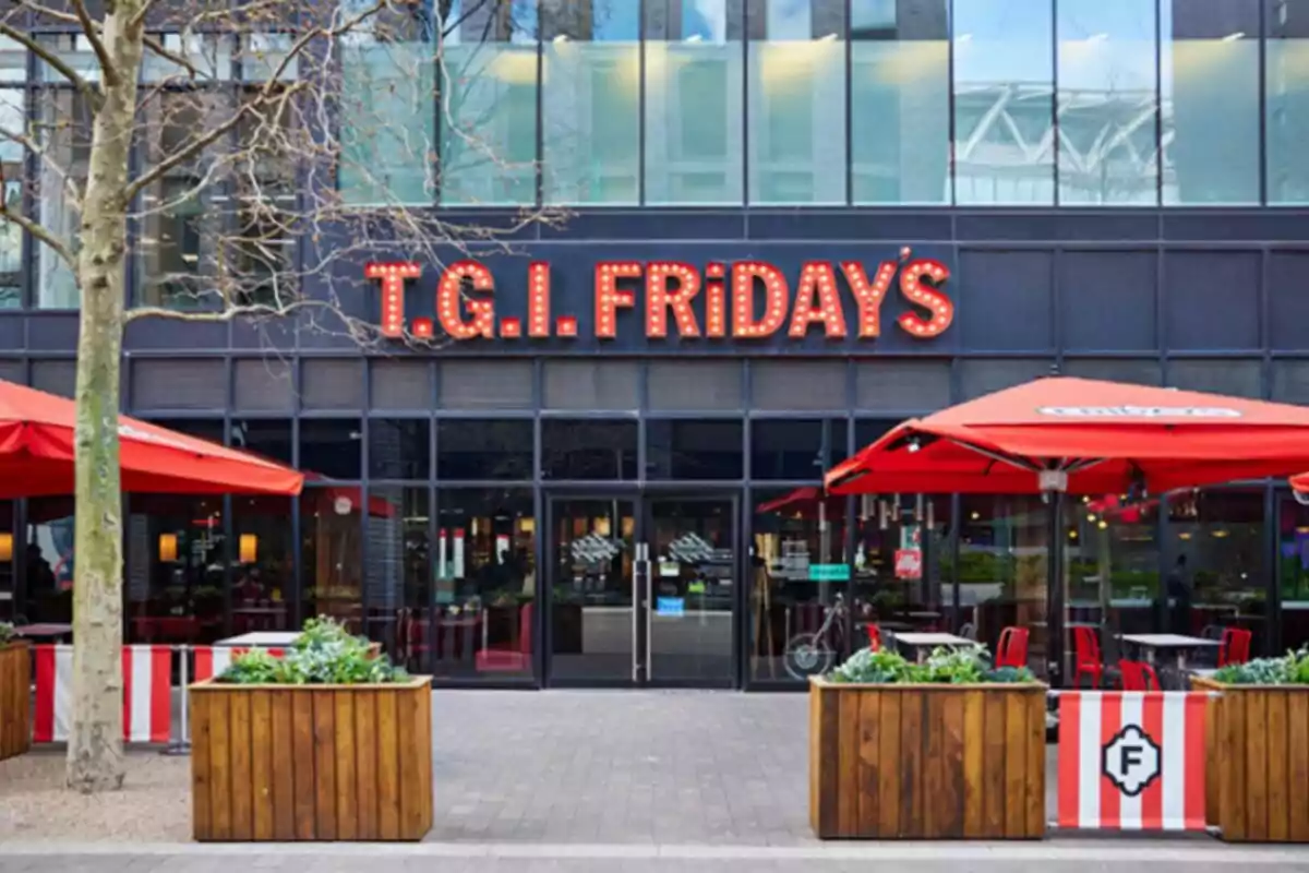 La imagen muestra la fachada de un restaurante T.G.I. Fridays con un letrero iluminado y sombrillas rojas en el área exterior. La imagen muestra la fachada de un restaurante T.G.I. Fridays con un letrero iluminado y sombrillas rojas en el área exterior.