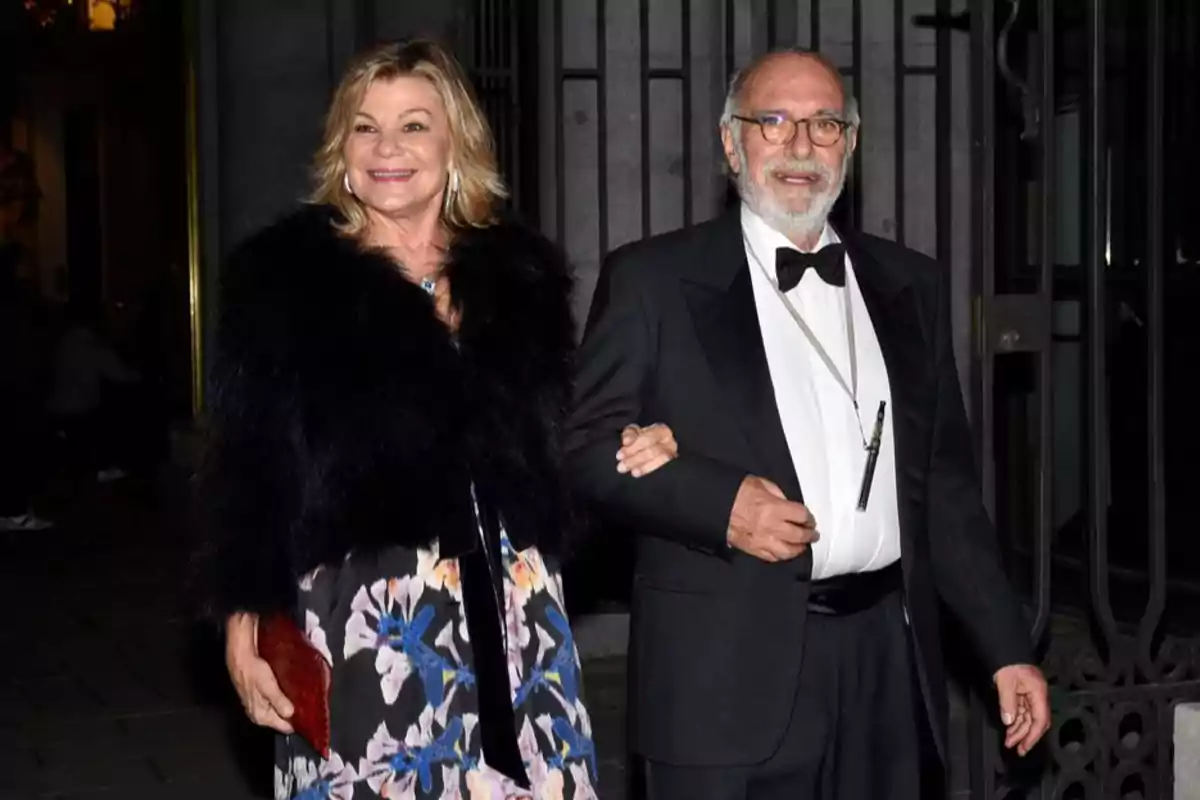 Una pareja elegantemente vestida, con la mujer usando un abrigo de piel y un vestido floral, y el hombre con un esmoquin y pajarita, posando juntos y sonriendo frente a una entrada con rejas.