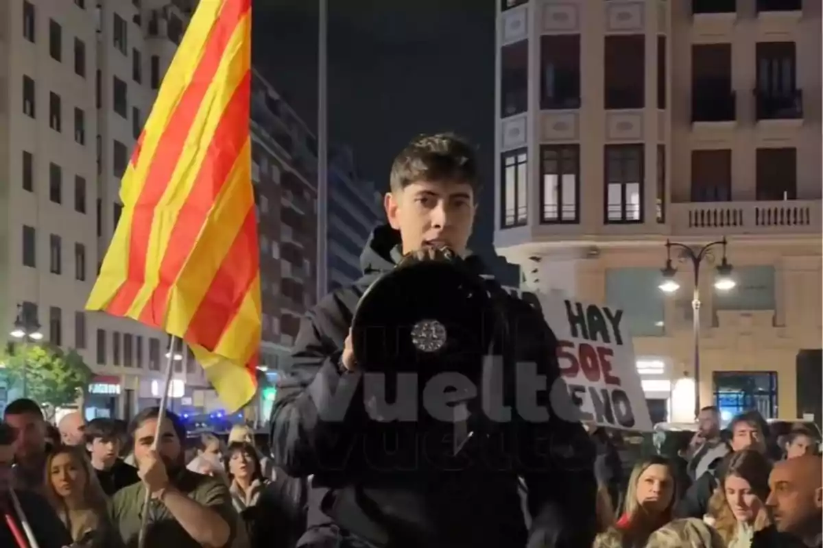 Una persona sostiene un megáfono en una manifestación nocturna con una bandera de rayas rojas y amarillas y un cartel que dice "HAY 50€ NO" en el fondo.