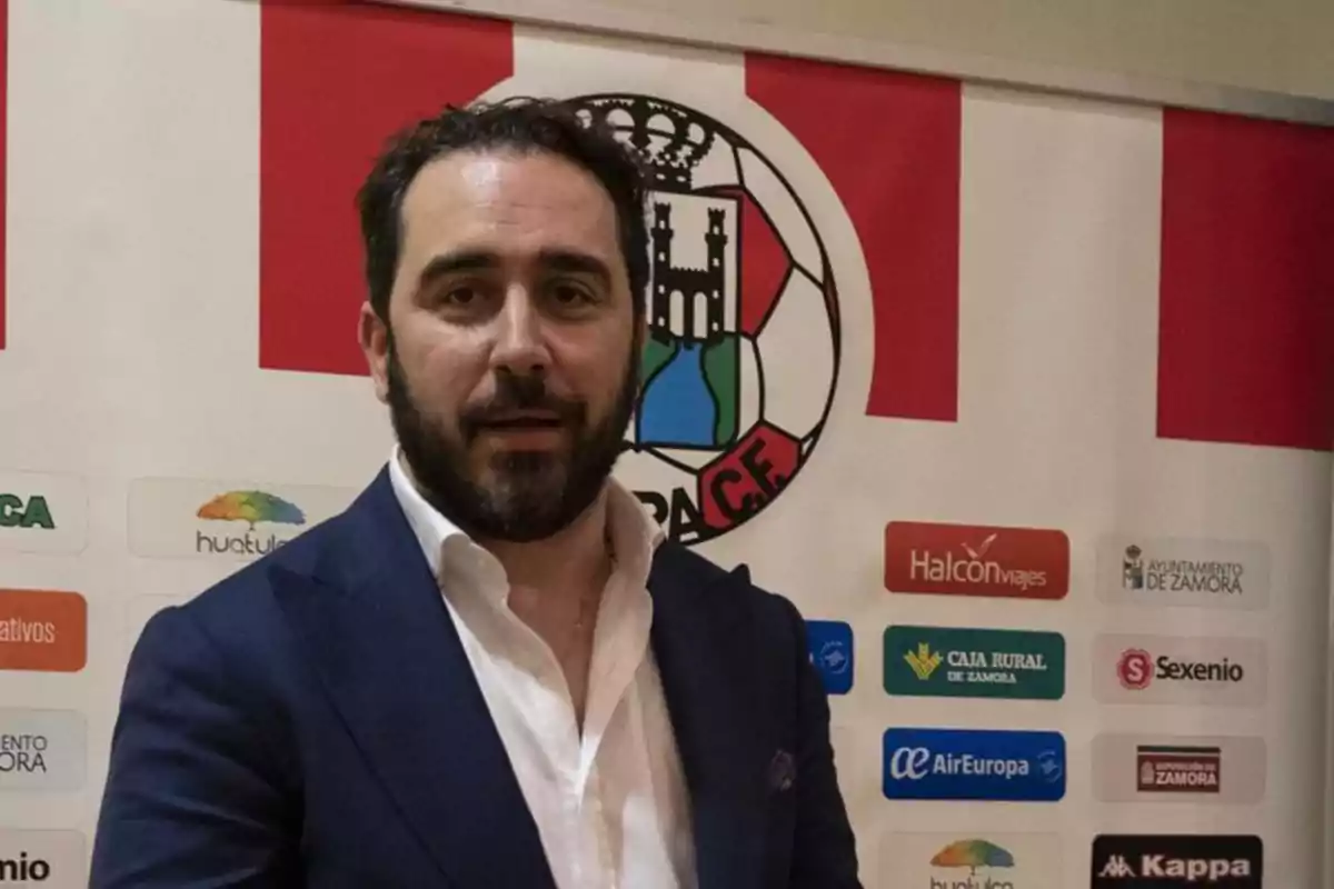 Hombre con barba y traje azul posando frente a un cartel con el escudo de un club de fútbol y varios logotipos de patrocinadores Hombre con barba y traje azul posando frente a un cartel con el escudo de un club de fútbol y varios logotipos de patrocinadores