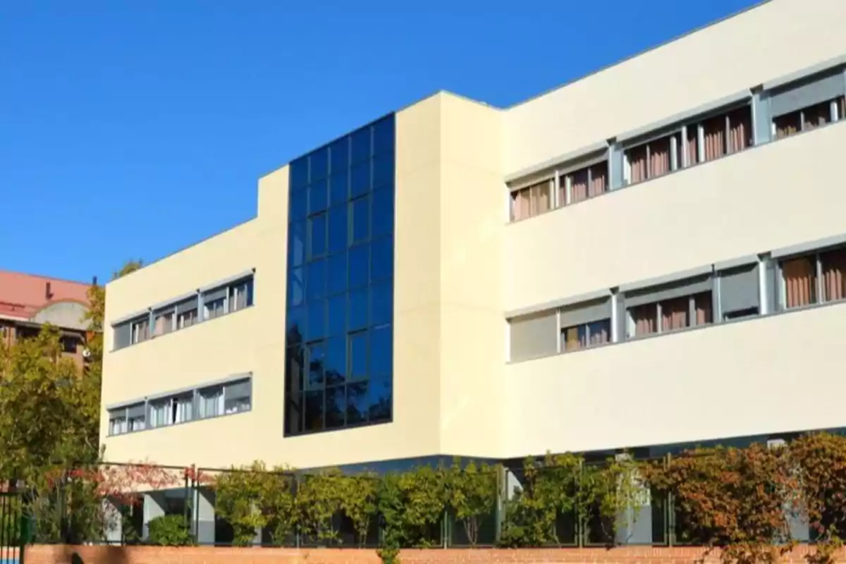 Edificio moderno de tres pisos con fachada clara y ventanales grandes rodeado de vegetación bajo un cielo despejado