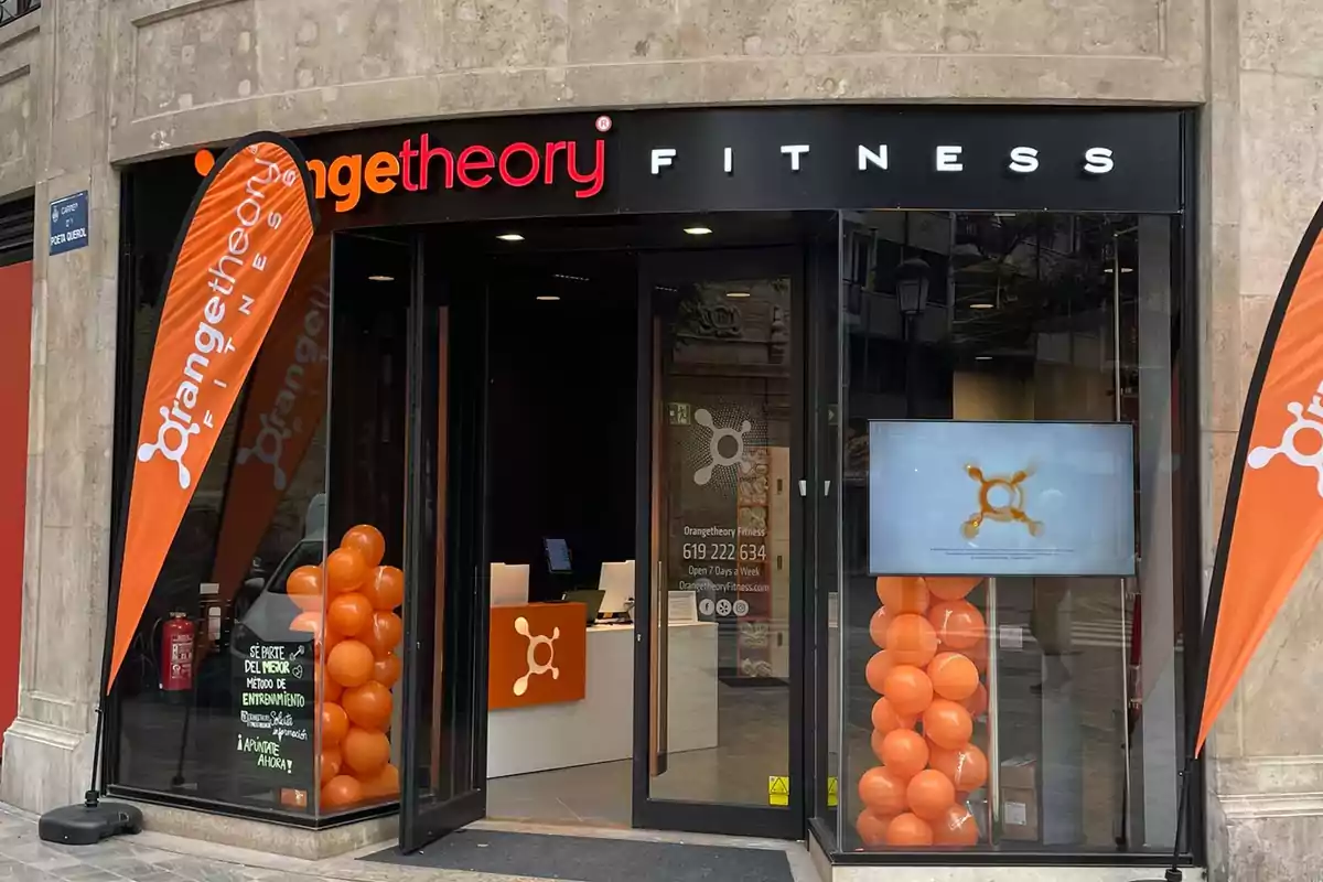 Entrada de un gimnasio Orangetheory Fitness con banderas y globos naranjas en la puerta.