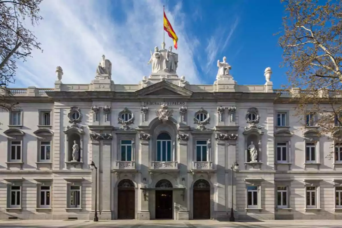 Edificio histórico con fachada neoclásica y una bandera ondeando en la parte superior, rodeado de árboles y bajo un cielo azul. Edificio histórico con fachada neoclásica y una bandera ondeando en la parte superior, rodeado de árboles y bajo un cielo azul.