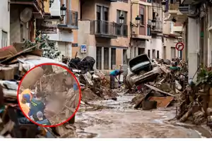 Una calle llena de escombros y un coche dañado tras una inundación, con personas trabajando en la limpieza.