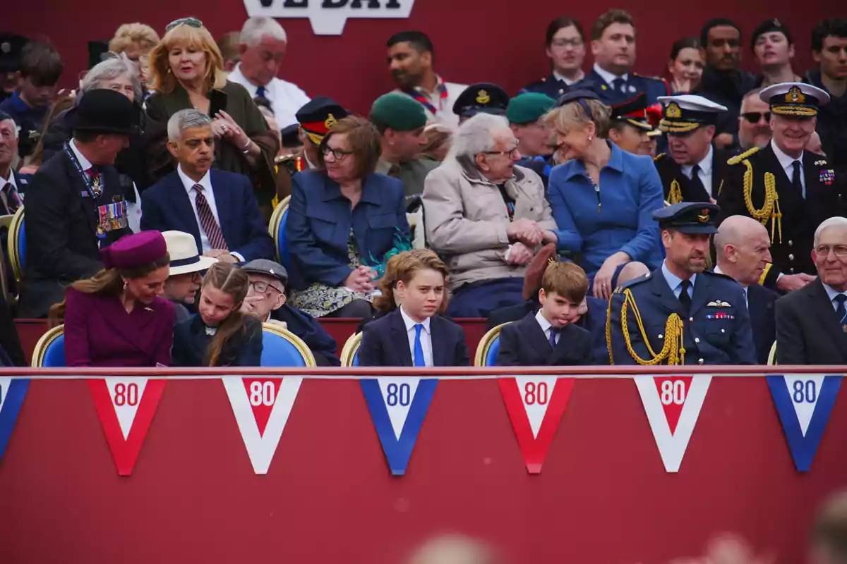 Un grupo de personas, algunas con uniformes militares, están sentadas en una tribuna decorada con banderines rojos, blancos y azules con el número 80, mientras observan un evento. Un grupo de personas, algunas con uniformes militares, están sentadas en una tribuna decorada con banderines rojos, blancos y azules con el número 80, mientras observan un evento.