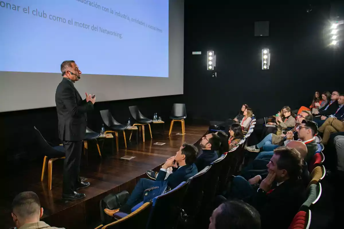 Un hombre dando una presentación en un auditorio frente a una audiencia atenta, con sillas vacías en el escenario y una pantalla proyectando texto.