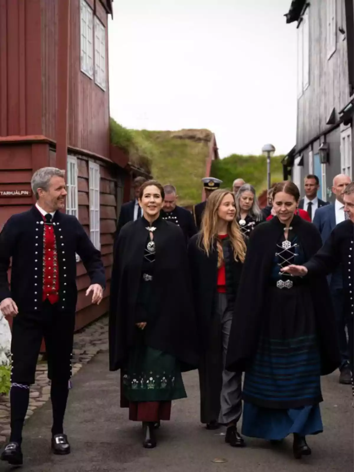 Grupo de personas caminando por una calle entre casas de madera, algunas de ellas llevan trajes tradicionales y capas oscuras Grupo de personas caminando por una calle entre casas de madera, algunas de ellas llevan trajes tradicionales y capas oscuras