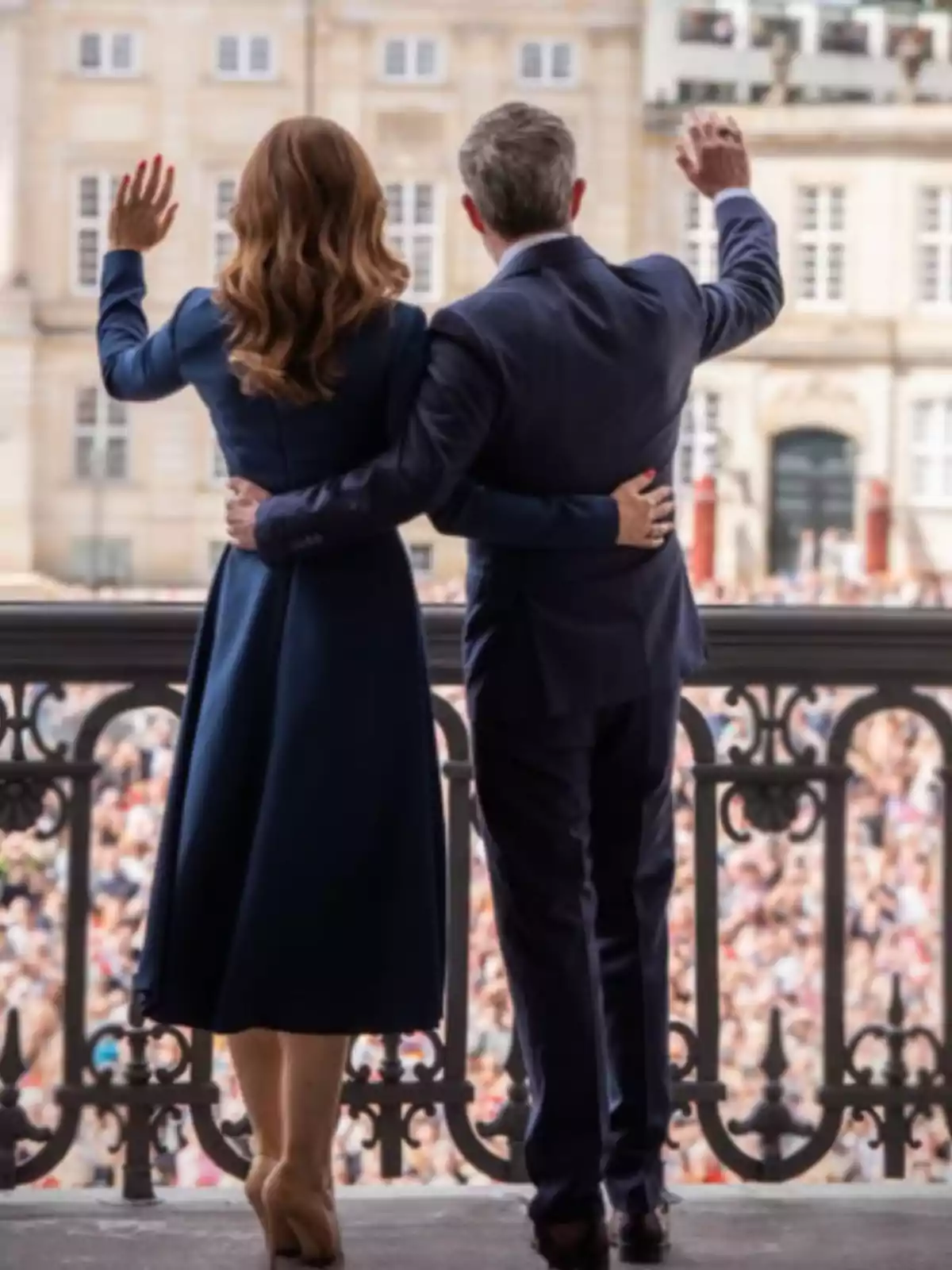 An elegantly dressed couple greets a crowd from a balcony. An elegantly dressed couple greets a crowd from a balcony.
