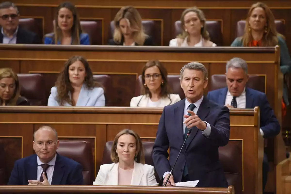 Un hombre de traje hablando en un parlamento con varias personas sentadas detrás de él.