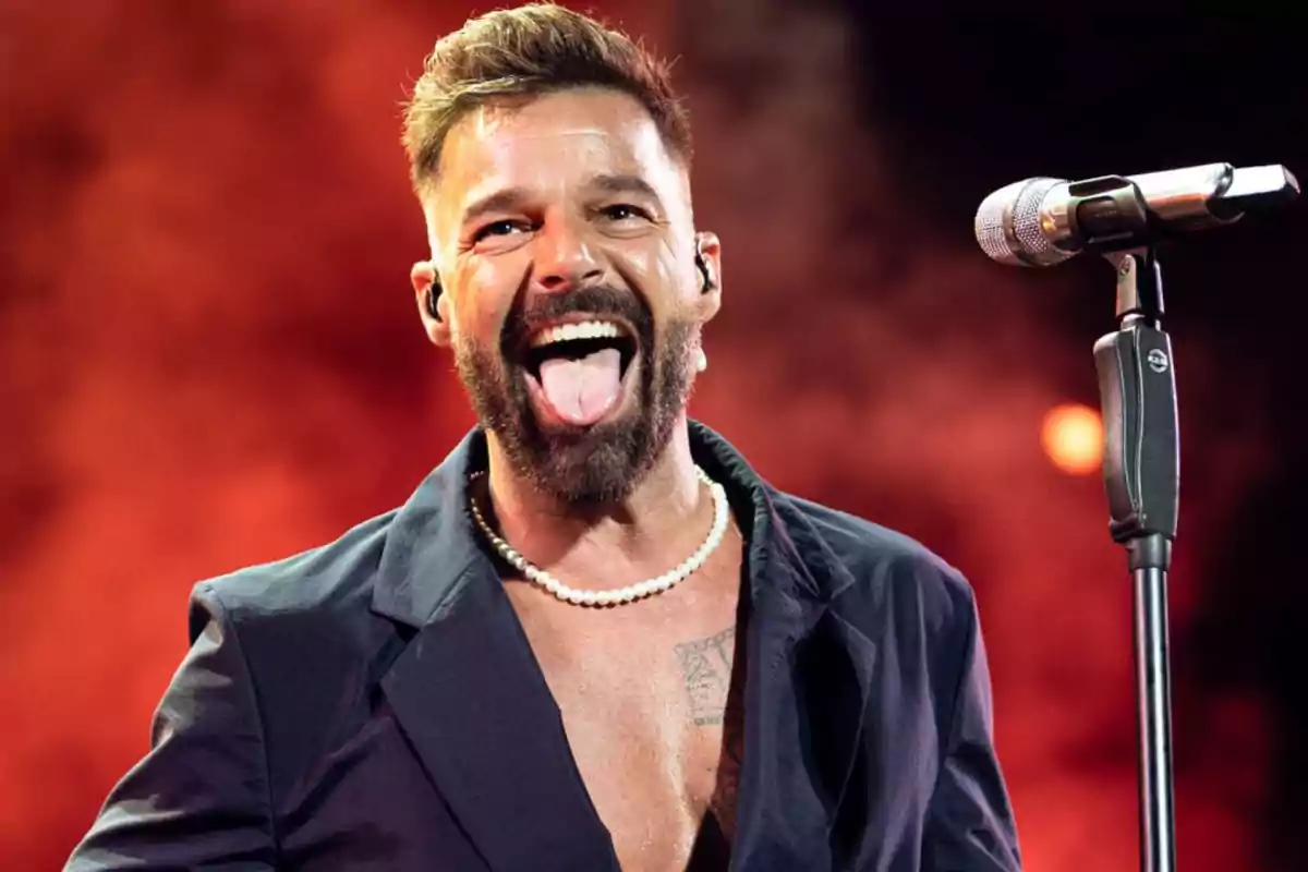 Persona con barba y collar de perlas sacando la lengua frente a un micrófono con fondo rojo.