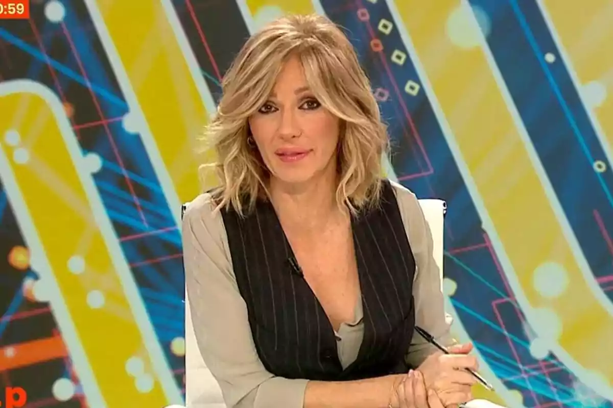 Una persona con cabello rubio y ondulado está sentada frente a un fondo colorido y geométrico, vistiendo una blusa clara y un chaleco oscuro. Una persona con cabello rubio y ondulado está sentada frente a un fondo colorido y geométrico, vistiendo una blusa clara y un chaleco oscuro.