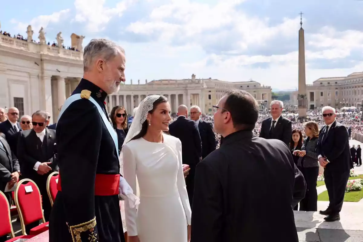 Felipe and Letizia elegantly dressed are conversing with a priest in a crowded square with classical architecture in the background. Felipe and Letizia elegantly dressed are conversing with a priest in a crowded square with classical architecture in the background.