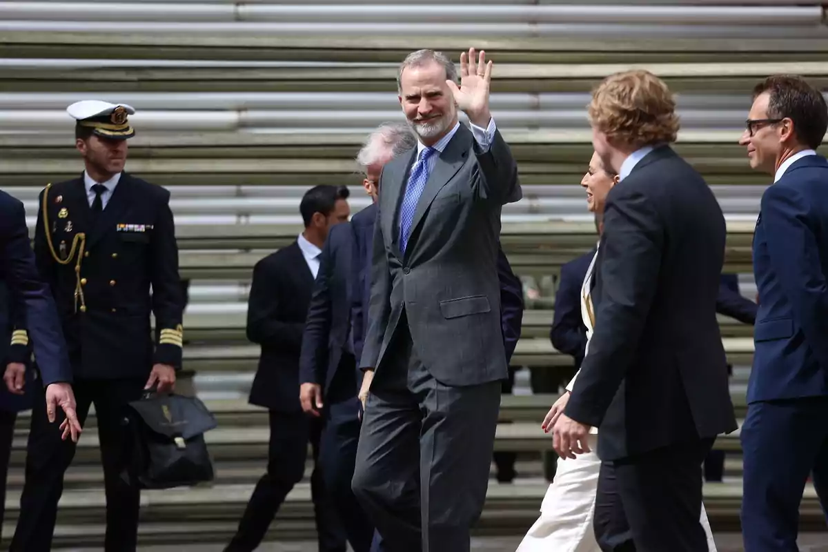 Felipe VI con traje gris y corbata azul saluda con la mano levantada mientras camina acompañado de varias personas vestidas formalmente, incluyendo un oficial naval. Felipe VI con traje gris y corbata azul saluda con la mano levantada mientras camina acompañado de varias personas vestidas formalmente, incluyendo un oficial naval.