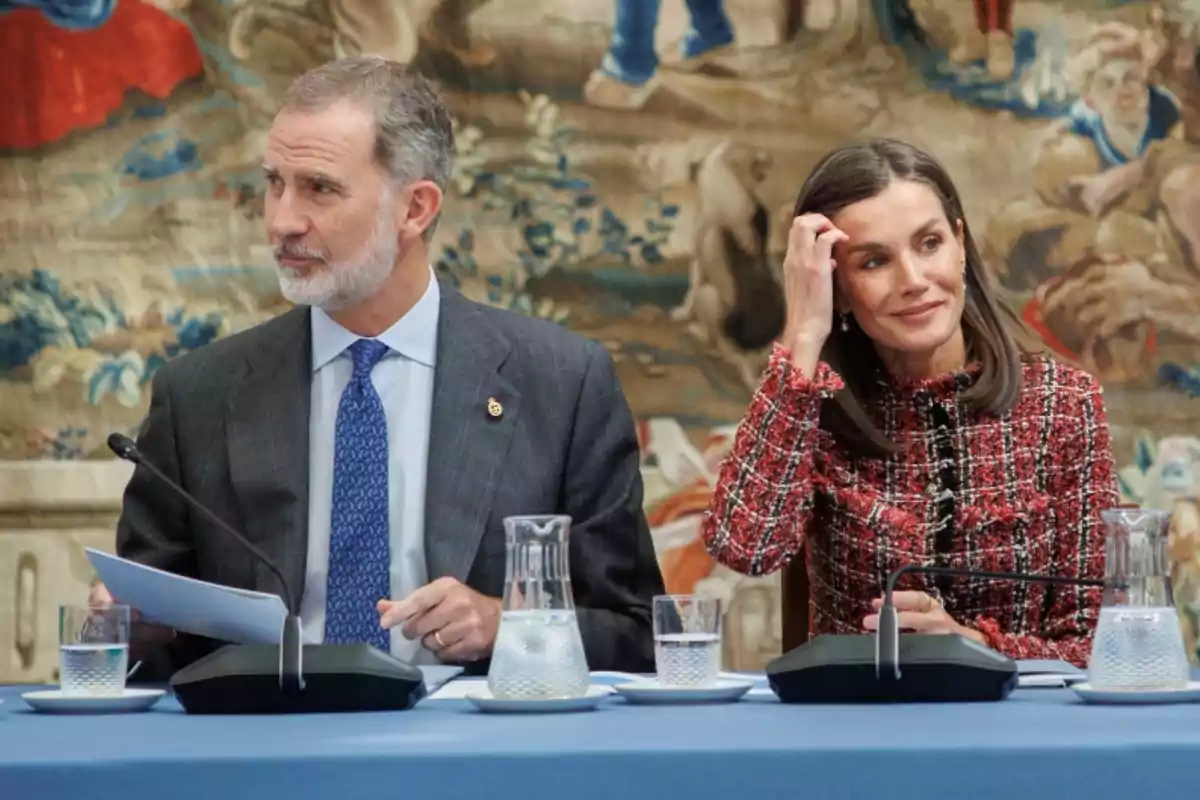 El rey Felipe VI y la reina Letizia de España, sentados juntos en una mesa, participan en un evento oficial. Felipe VI, con traje oscuro y corbata azul, sostiene unos documentos mientras Letizia, vestida con un abrigo de tweed rojo, se toca el cabello y sonríe. En el fondo, un tapiz decorativo con motivos históricos