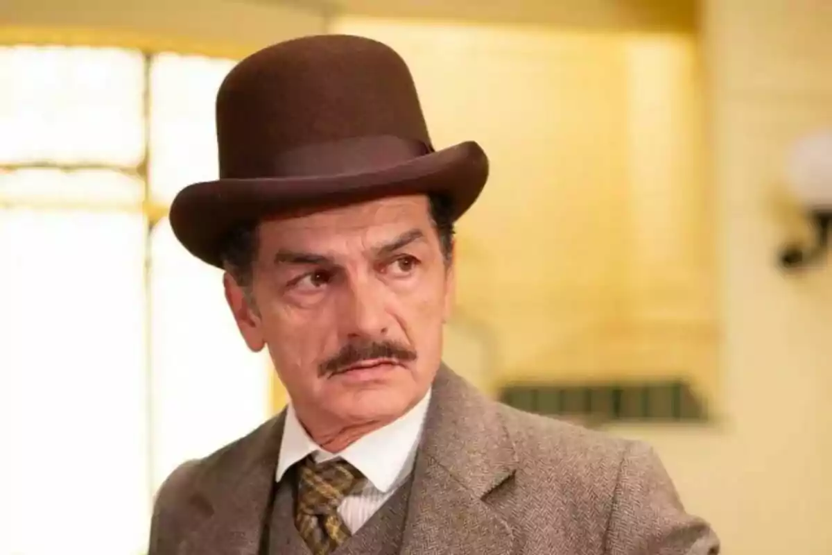 Carles Sanjaime as Fermín Villanueva with moustache and brown top hat, dressed in brown suit and checkered tie, looking left in a lighted interior setting.