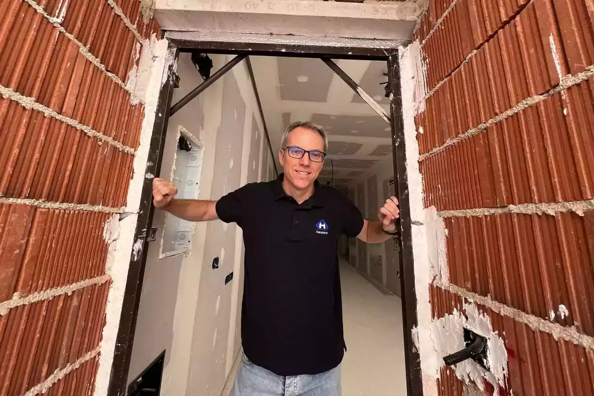 Un hombre con gafas y camiseta negra sonríe mientras se apoya en el marco de una puerta en construcción.