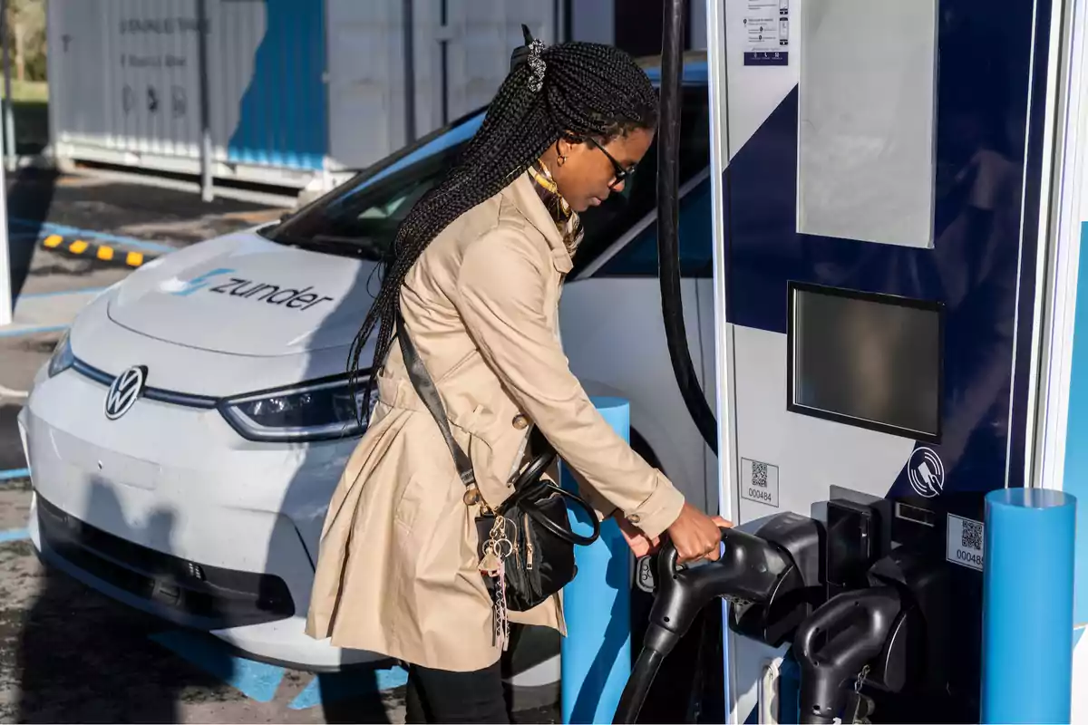 Una mujer con abrigo beige y gafas de sol está cargando un coche eléctrico en una estación de carga, con un vehículo Volkswagen blanco de fondo.